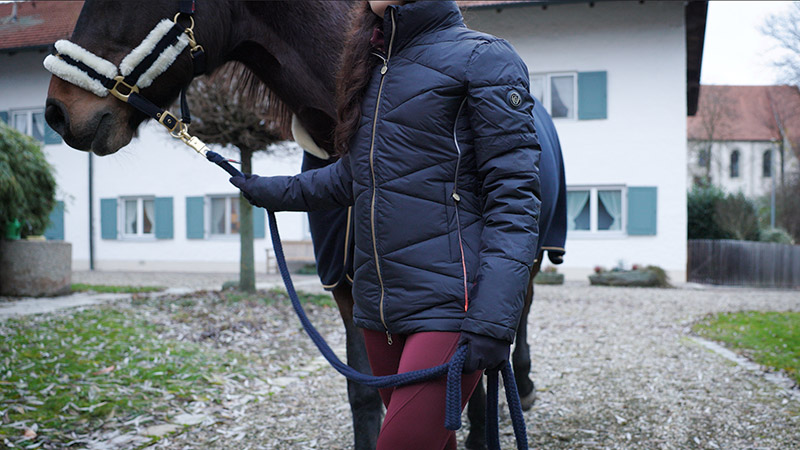 Frau in Covalliero Reitkleidung und LED Steppjacke führt Pferd über den Hof