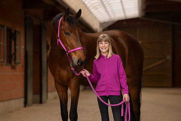 Ein Mädchen in pinkfarbener Reitjacke führt ein braunes Pferd mit pinkem Halfter im Stall